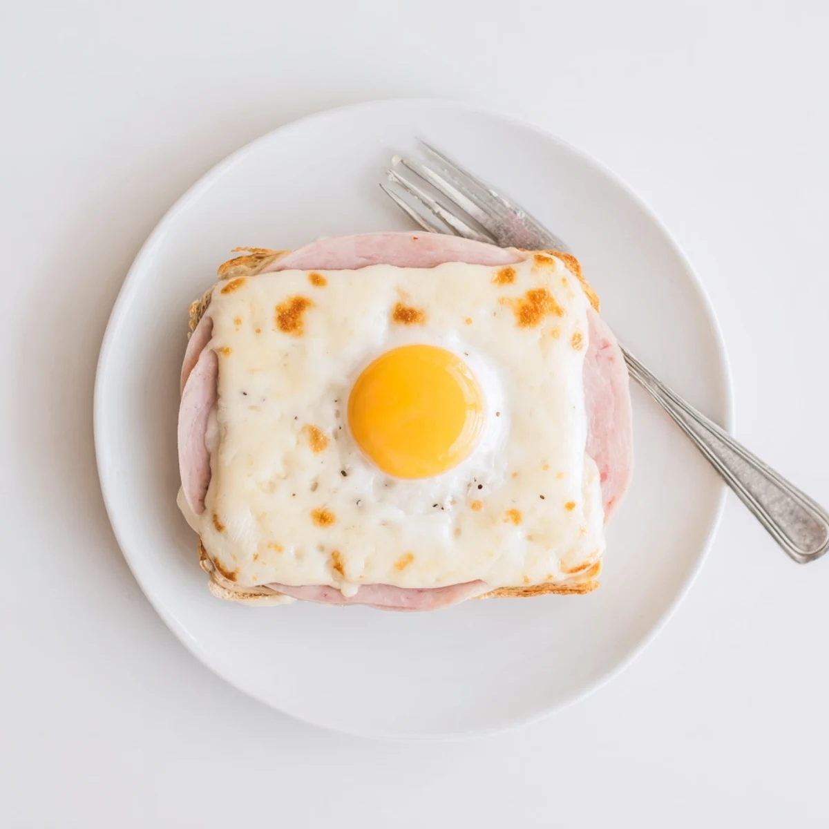 A golden Croque Madame sandwich topped with a sunny-side-up egg on a rustic plate, béchamel dripping down the sides.  