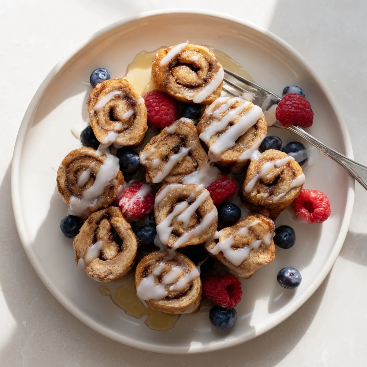 Golden brown Cinnamon Roll French Toast Bites glistening with drizzled icing, resting on a white plate with fresh berries.