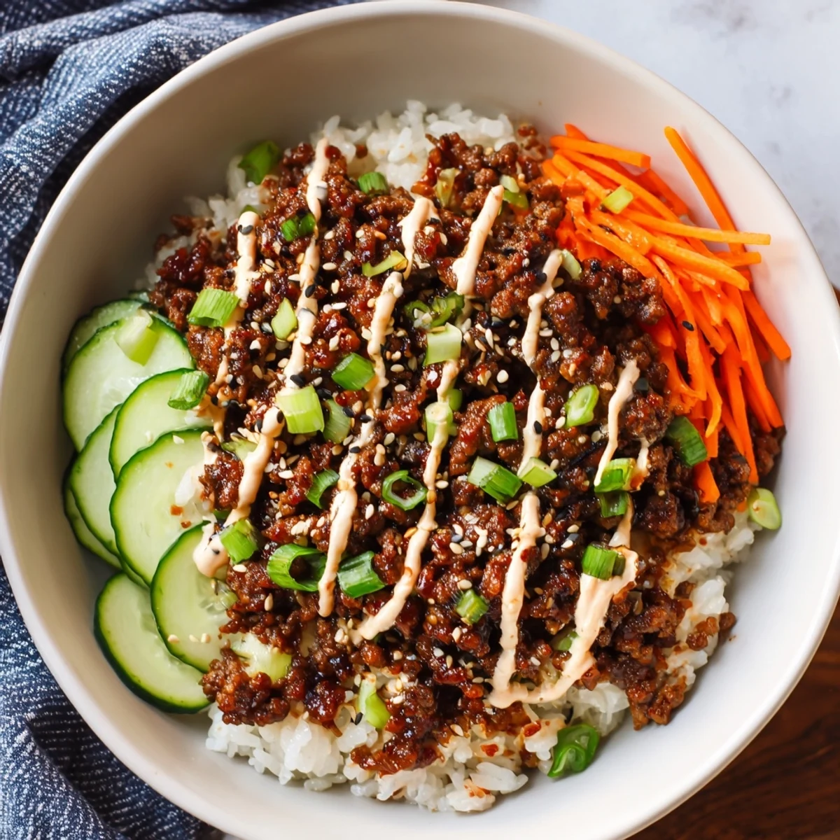 A vibrant Korean Beef Bowl with Spicy Mayo sits atop fluffy white rice, garnished with crunchy carrots, cucumbers, and sesame seeds.
